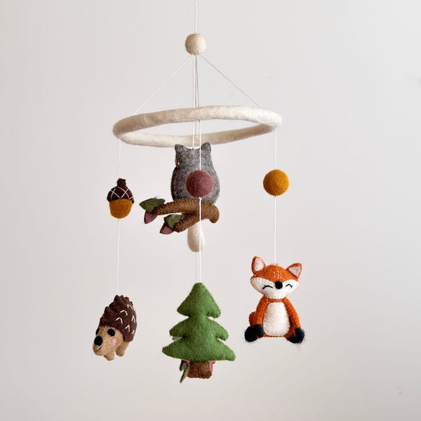 A handcrafted wool felt baby mobile, featuring whimsical forest characters, hangs from a white string against a light background. The mobile showcases an owl, fox, mushroom, hedgehog, acorn, and pine tree, crafted with simple shapes and natural colors in shades of brown, green, and beige. Each character is carefully designed to create a warm and inviting atmosphere suitable for a baby's room.