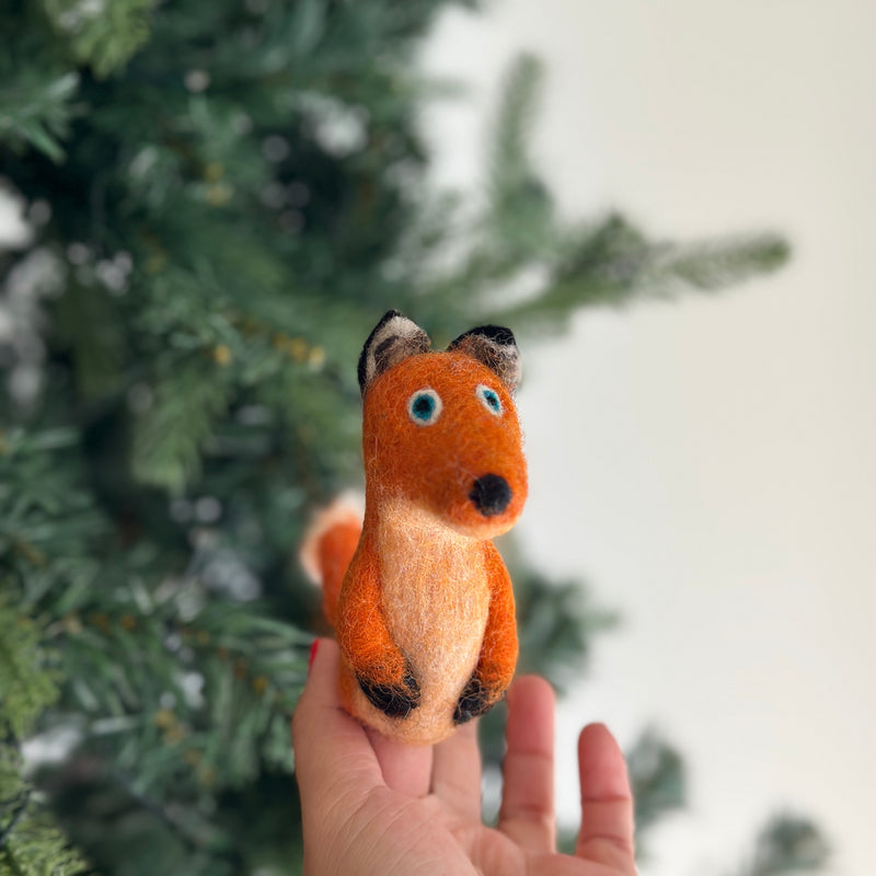 A vibrant orange wool felt fox finger puppet is held upright in a hand, set against a blurred Christmas tree background. This charming, fair-trade handmade fox puppet features a lighter chest, black accents on its paws, nose, and ear backs. Its small blue eyes and round tail enhance its cute, whimsical appeal, making it a unique handcrafted felt kids toy.