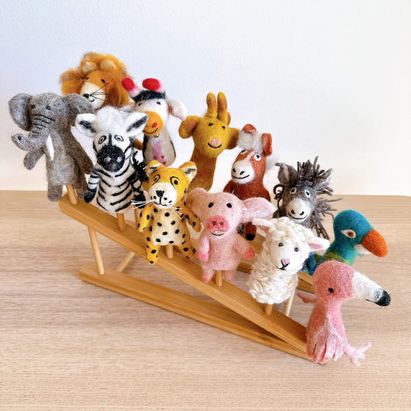 A colorful collection of wool felt finger puppets, featuring diverse animals like elephants, giraffes, and zebras, are arranged on a wooden shelf. The playful display showcases the unique designs and vibrant colors of each puppet, with some even holding hands or interacting with each other. This set of 12 handmade wool felt finger puppets is made from 100% wool and includes a bamboo shelf for organization and a soft cotton drawstring bag for protection.