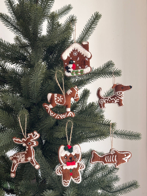 A set of six cheerful needle felted gingerbread ornaments hangs on a lush green Christmas tree, set against a soft off-white background. Each handmade ornament, suspended by a jute string, features unique designs: a gingerbread house with a snowy roof, a rocking horse, a dog with white swirls, a reindeer with snowflakes, a cat with a red bow and holly, and a shark with white accents.