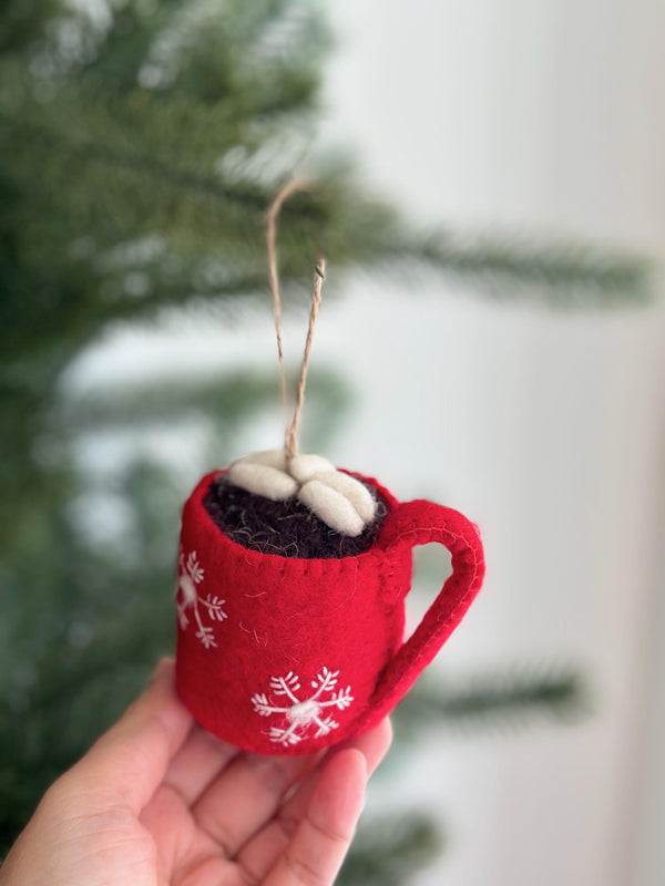 A handmade hot cocoa marshmallow felt ornament is held by a female hand, presented against a blurred green Christmas tree and a light wall, creating a warm, festive mood. This unique Christmas tree ornament is shaped like a red mug with stitched white snowflakes, filled with dark brown cocoa and white marshmallows. A jute string is attached for hanging.