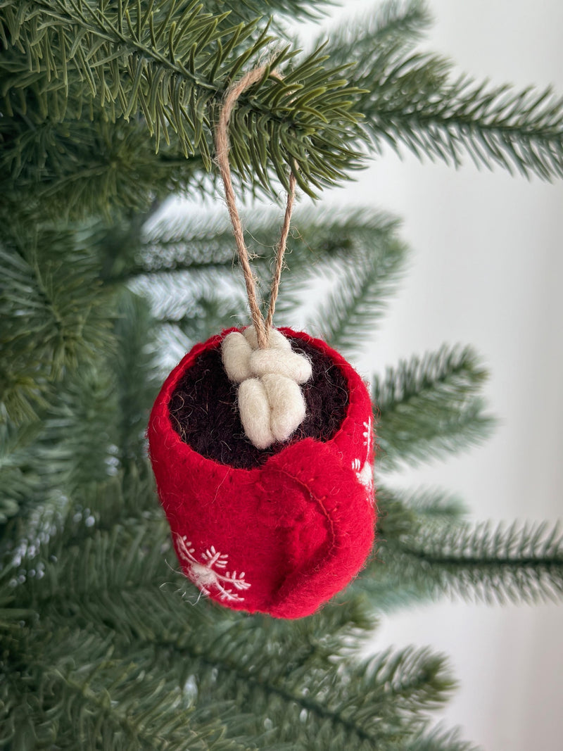 A handmade felt hot cocoa marshmallow ornament hangs from a green Christmas tree branch, set against a soft white background. The unique Christmas ornament features a red cup with white snowflake patterns, filled with brown felt cocoa and topped with fluffy white felted marshmallows. A jute string loop is attached for hanging, completing this charming Christmas tree decoration.