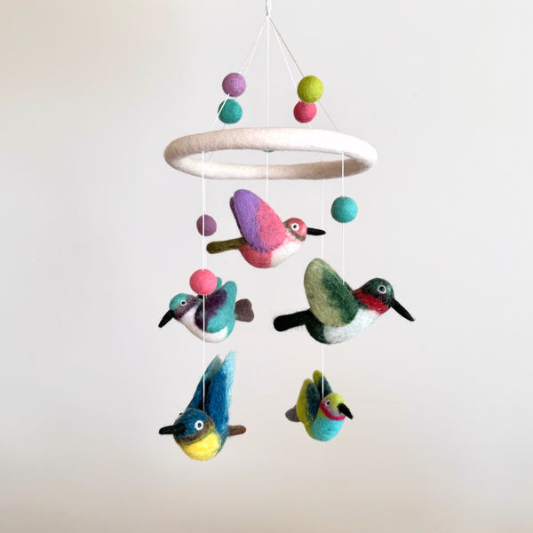 A charming Hummingbird Baby Mobile hangs from the ceiling, suspended by a string against a white background. The mobile features five colorful hummingbirds with unique wing positions on a white felt frame, creating a lively display.