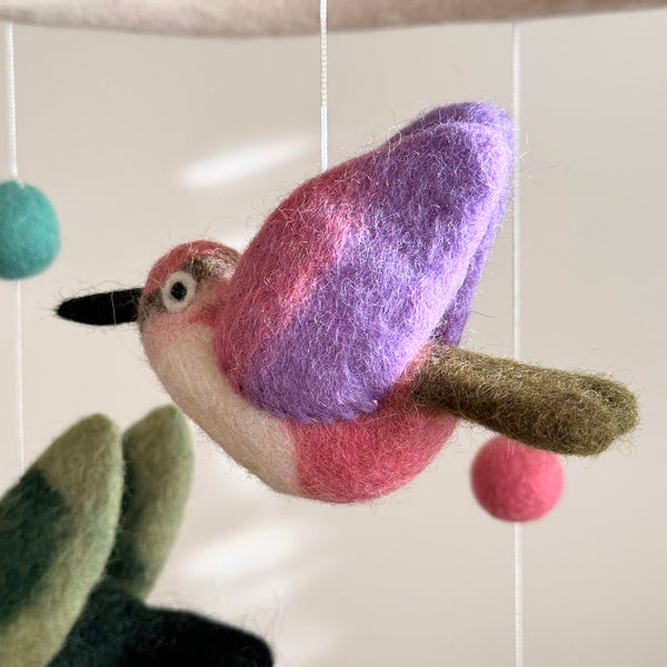 A handcrafted baby mobile featuring five colorful hummingbirds in various poses, hanging from a string attached to a white felt frame. The background is a wooden shelf with other items, creating a warm and inviting atmosphere.