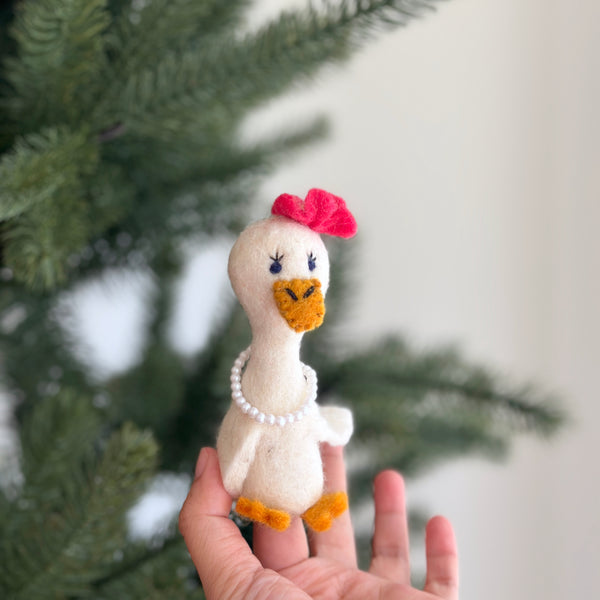 A hand holding a small, plush lady duck finger puppet with a red bow on its head, featuring a yellow beak and feet, wearing a pearl necklace. The background is blurred but suggests a cozy indoor setting, possibly in front of a Christmas tree decorated with pine cones and ornaments.