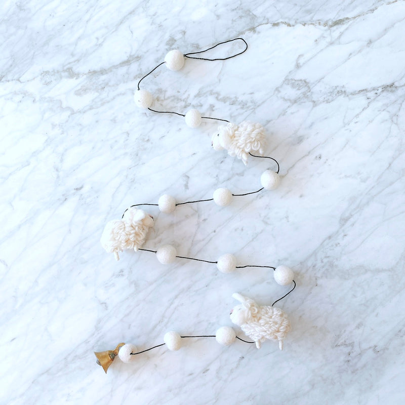 A Mini Sheep felt garland is displayed on a white marble surface. The garland features three small, fluffy white felt sheep, each with a skillfully crafted 3D design, evenly spaced along a thin black string, interspersed with white felt balls. A copper bell dangles at one end, adding a sweet, whimsical accent.