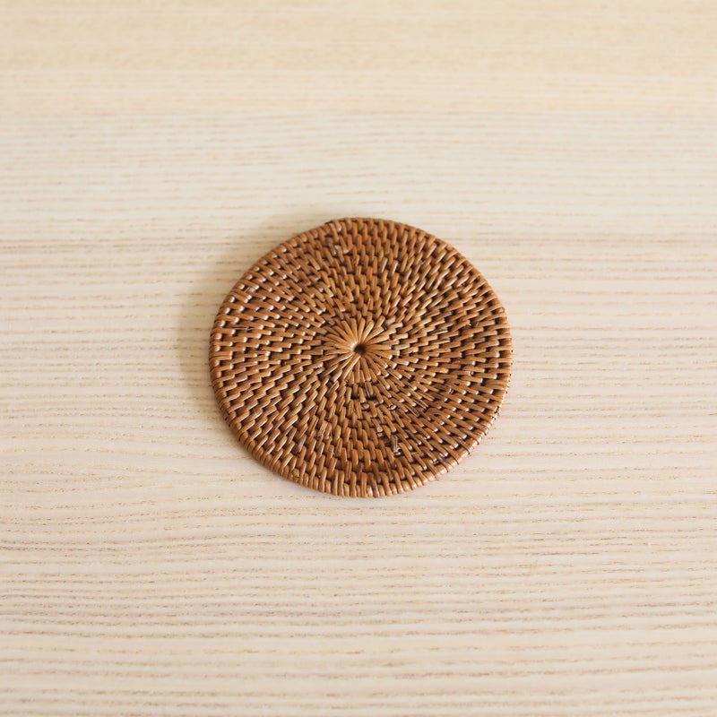 Handcrafted Bali rattan coaster, one from a set of 5 handmade in Bali, is beautifully displayed in a minimalist top-down shot. The single, golden-brown coaster is centered on a light, fine-grained wood surface, its intricate sunburst weave creating a striking textural focal point. The clean, simple environment enhances the coaster's natural, artisanal feel, evoking a sense of calm and organic elegance.