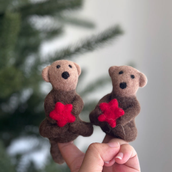 A handcrafted wool felt sea otter finger puppet, holding a bright red sea star, is shown on a female hand against a blurred Christmas pine tree background. This charming puppet features soft brown wool felt in varying shades, with a lighter head and darker body. It has small black eyes and a nose, exuding a cozy, playful spirit.