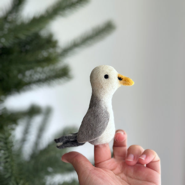 A felt seagull finger puppet resting against a Christmas tree, its grayish-blue body, white wings, yellow beak, and black eyes visible. The seagull felt finger puppet is made of wool felt, with intricate details that evoke the spirit of the coastal bird.