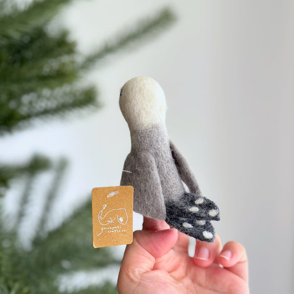 A hand holds a gray felt seagull finger puppet with white markings on its wings, tail, and head. The puppet features detailed beak, eyes, and feet made from the same felt material. This handmade wool felt seagull finger puppet is crafted in Nepal by female artisans, promoting fair-trade and environmentally friendly practices.
