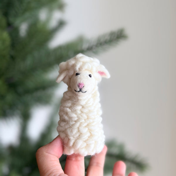 A hand holds a small, wool felt finger puppet sheep on a white background. The sheep has a fluffy texture and black details for its eyes, nose, and mouth, giving it a friendly appearance.