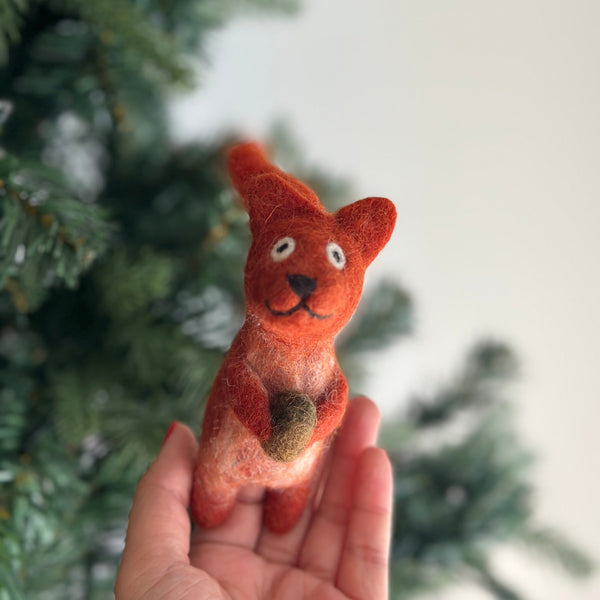 A handmade wool felt squirrel with acorn finger puppet is gently held by a female hand, set against a softly blurred Christmas tree background. This charming orange puppet features a dark brown acorn in its paws, white eyes, a stitched smile, and perky ears, giving it a curious and playful expression.