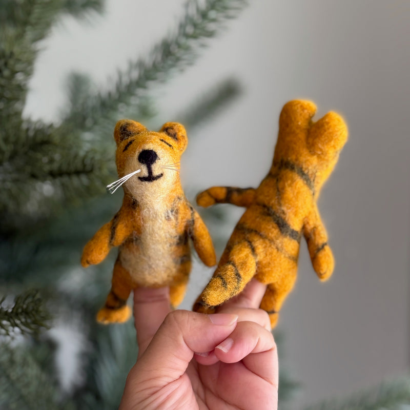 A yellow and orange wool felt tiger finger puppet is presented by a female hand against a blurred, festive backdrop, possibly near a Christmas tree. This handmade needle felted tiger finger puppet features black stripes, a stitched smile, and whiskers, while another tiger puppet nearby shows its striped back. Crafted from 100% wool felt, these soft 3D tiger puppets are designed for interactive play, highlighting their handcrafted quality and whimsical appeal.