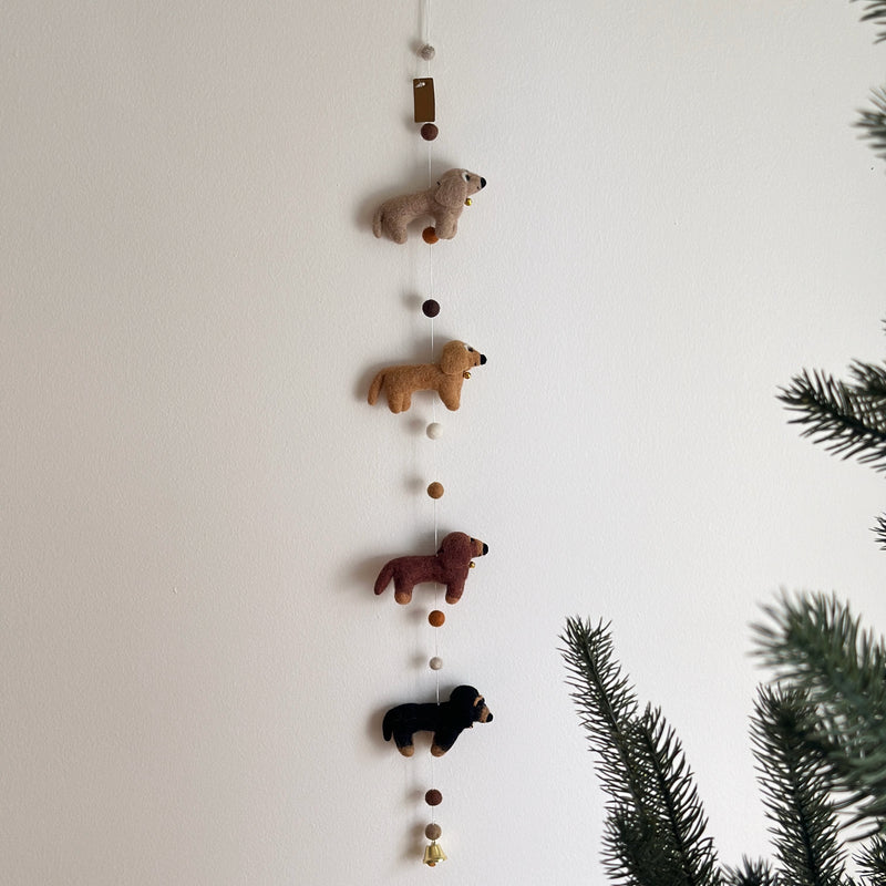 A felt Dachshund dog garland hangs on a plain white wall, adding a playful and festive touch. It features four evenly spaced felt Dachshunds, each in a different color: cream, khaki, brown, and black. A blurred evergreen branch is visible on the right side.