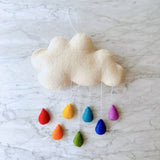 A whimsical wool felt garland, suspended from a string against a marble countertop, features a fluffy white cloud adorned with colorful rainbow raindrops. The mobile is handcrafted and adds a playful touch to any child's room.