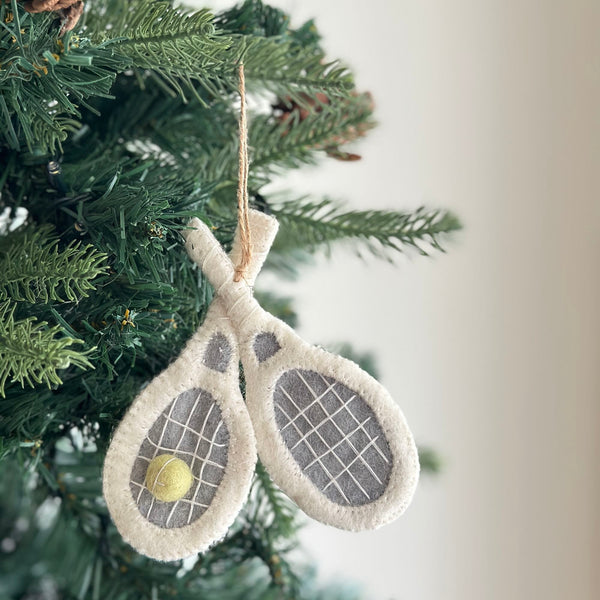 A felt tennis racket ornament hangs on a Christmas tree, adding a playful and sporty touch. This unique Christmas ornament features two white tennis rackets crossing, with white borders and gray netting. A yellow tennis ball is nestled centrally between them. Suspended by a jute string, the handcrafted felt ornament blends seamlessly with the festive green tree adorned with a brown pine cone.