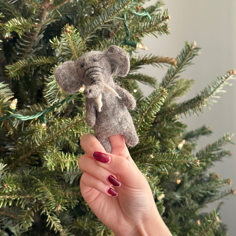 A handmade wool felt elephant finger puppet is gently held by a female hand with burgundy nail polish, set against a festive Christmas tree adorned with subtle lights. This grey elephant finger puppet, crafted from wool felt with 3D needle felting, features floppy ears, tusks, and a wire in its trunk for added dimension.