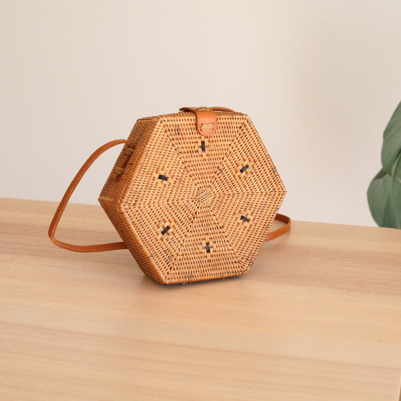A woven rattan crossbody bag resting on a wooden surface, featuring a hexagonal shape with a honeycomb pattern design. The bag has an adjustable brown leather strap and is adorned with a unique Batik lining. The overall atmosphere suggests a casual setting, possibly in a home or boutique store.