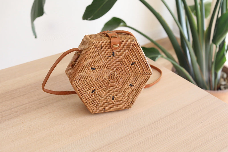 A Bali Hexagon Rattan Crossbody Bag sits on a wooden surface, showcasing its natural rattan Ata grass construction and unique hexagonal shape. The bag features a secure snap closure and adjustable leather strap, with a batik lining visible through the open top. This eco-friendly, handcrafted piece is perfect for adding bohemian charm to any outfit.