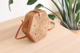 A Bali Hexagon Rattan Crossbody Bag rests on a wooden surface, showcasing its natural rattan Ata grass texture and unique hexagonal shape. The bag features a batik lining and secure snap closure, with a real leather strap that is adjustable.