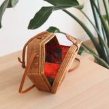 A Bali Hexagon Rattan Crossbody Bag rests on a wooden surface, showcasing its unique hexagonal shape. The bag is made from natural rattan Ata grass with a visible woven texture and features a real leather strap with an adjustable buckle. A batik print design adorns the interior.