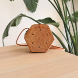 A small, woven hexagonal purse sits on a wooden surface, featuring a brown leather strap and batik lining visible through the weave. The background shows a plant and a portion of a white wall, reflecting a casual and bohemian style.