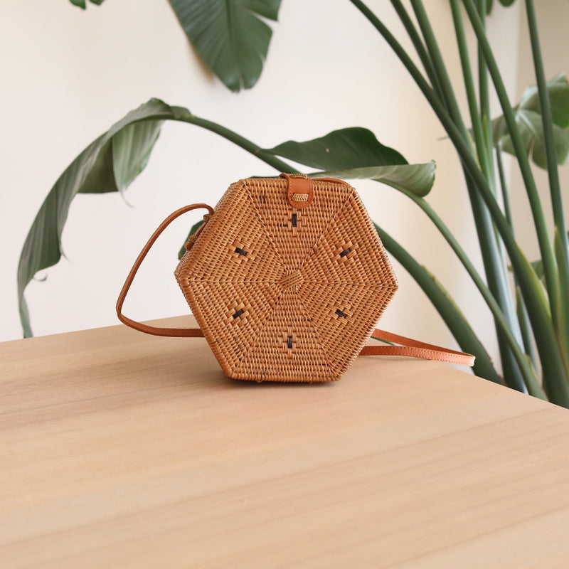 A small, woven hexagonal purse sits on a wooden surface, featuring a brown leather strap and batik lining visible through the weave. The background shows a plant and a portion of a white wall, reflecting a casual and bohemian style.