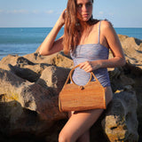 A stylish woman is holding a Bali Rattan Wheel Handbag handmade by Ganapati Crafts Co. in Bali standing on a beach in Bali where Bali rattan bags are made