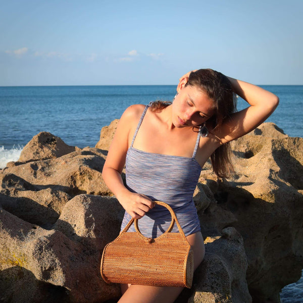 A stylish woman is holding a Bali Rattan Wheel Handbag handmade by Ganapati Crafts Co. in Bali standing on a beach in Bali where Bali rattan bags are made