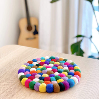 A cheerful, circular rainbow wool felt pot trivet, handcrafted from organic lamb wool felt balls, rests on a light wooden table. This approximately 8-inch diameter trivet features tightly packed, hand-stitched felt in white, yellow, blue, orange, and green, creating a vibrant, textured surface. A blurred guitar and potted plant are visible in the background, adding a domestic touch.