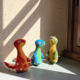 The Dino Brothers Brontosaurus wool felt finger puppets stand on a sunlit surface near a window, creating a playful scene. The trio includes a red and orange puppet with white spots, a blue and green one with white spots, and a green and yellow puppet also with white spots. Each handmade, needle felted puppet features a long neck and a slightly fuzzy texture from its 100% wool felt construction.