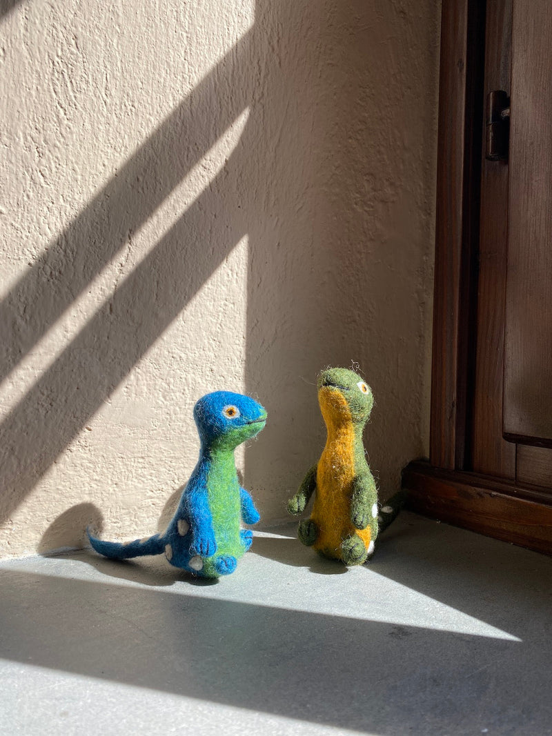 Two handcrafted wool felt Brontosaurus finger puppets, resembling the 'Dino Brothers', are displayed against a textured wall and a dark wood door frame in a warm, naturally lit setting. These needle-felted puppets, made from 100% wool, are positioned as if interacting playfully. One Brontosaurus finger puppet has a blue head and tail with a green, white-spotted body; the other features a green head, yellow body, and white spots, both showcasing long necks and 3D designs.