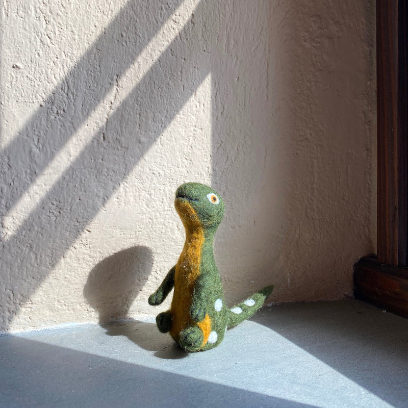 A handcrafted green wool felt Brontosaurus finger puppet sits in a sunlit corner, resting against a textured light beige wall with dark brown trim. This playful felted Brontosaurus puppet features a yellowish-tan neck and underbelly, with small white spots along its back and tail. Its needle felted design includes a long neck, small arms, and tiny round eyes, inviting imaginative play for kids.