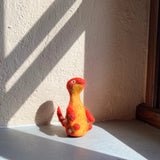 A vibrant wool felt Brontosaurus finger puppet from the Dino Brothers collection rests on a sun-drenched windowsill, with light streaming through window blinds. This handcrafted needle felted Brontosaurus toy features a lifelike 3D design with a long neck, small red felt limbs, and white body dots. Its body transitions with a beautiful color gradient from red at the top to yellow at the bottom, showcasing its artisanal quality.