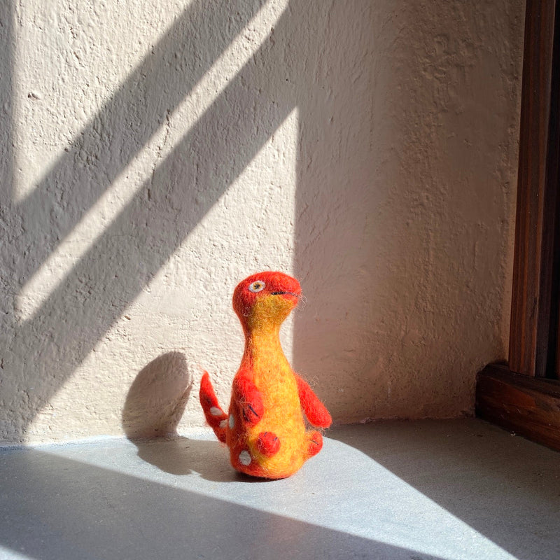 A vibrant wool felt Brontosaurus finger puppet from the Dino Brothers collection rests on a sun-drenched windowsill, with light streaming through window blinds. This handcrafted needle felted Brontosaurus toy features a lifelike 3D design with a long neck, small red felt limbs, and white body dots. Its body transitions with a beautiful color gradient from red at the top to yellow at the bottom, showcasing its artisanal quality.