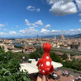 A vibrant red Brontosaurus wool felt finger puppet, part of the Dino Brothers collection, is prominently displayed on a person's finger. Crafted from 100% needle-felted wool, this whimsical dinosaur puppet features a long neck, majestic shape, and white polka dots. It stands in sharp focus against a blurred cityscape with buildings and bridges under a bright blue sky, suggesting a playful urban adventure.