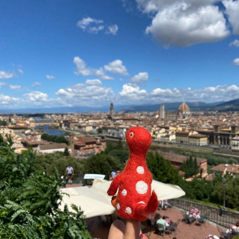 A vibrant red Brontosaurus wool felt finger puppet, part of the Dino Brothers collection, is prominently displayed on a person's finger. Crafted from 100% needle-felted wool, this whimsical dinosaur puppet features a long neck, majestic shape, and white polka dots. It stands in sharp focus against a blurred cityscape with buildings and bridges under a bright blue sky, suggesting a playful urban adventure.