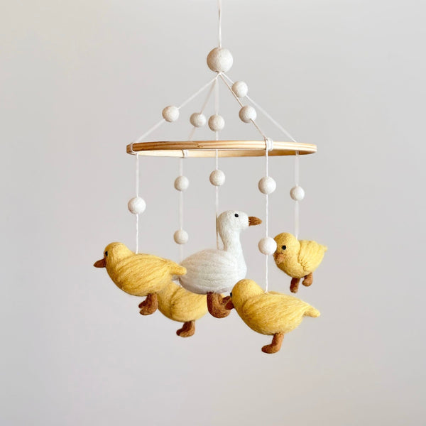 A felt baby mobile, featuring five white call ducks in various poses on a wooden frame, hangs from a ceiling. The mobile's neutral background showcases its playful design, with each duck having distinct expressions and details.
