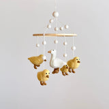 A felt baby mobile, featuring five white call ducks in various poses, hangs from a wooden hanger. The largest duck is at the center, surrounded by smaller ones, against a neutral background.