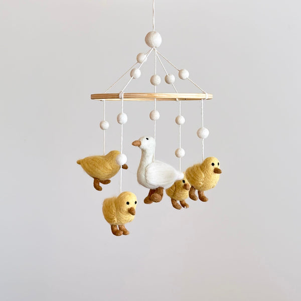 A felt baby mobile, featuring five white call ducks in various poses, hangs from a wooden hanger. The largest duck is at the center, surrounded by smaller ones, against a neutral background.