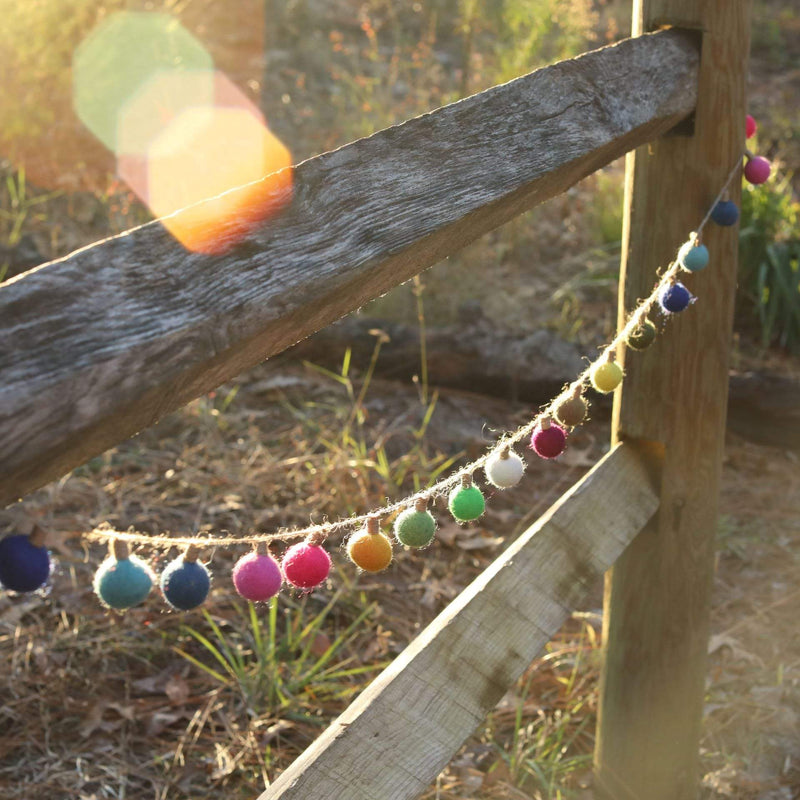 The Felt Color Ball Lightbulb Garland is draped across a rustic wooden fence in a sunlit outdoor setting. This cheerful wool felt garland features colorful, spherical balls resembling lightbulbs, crafted from 100% wool and 3D designed. The vibrant blue, teal, pink, red, yellow, and white balls are evenly spaced on a thin twine, adding a playful touch to the natural environment.