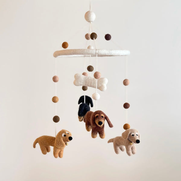 A colorful mobile featuring four adorable dachshunds, crafted from felt, hangs from the ceiling. The dachshunds are in various colors: cream, khaki, brown, and black. A small felt bone is attached at the center, adding a playful touch. The background is plain white.