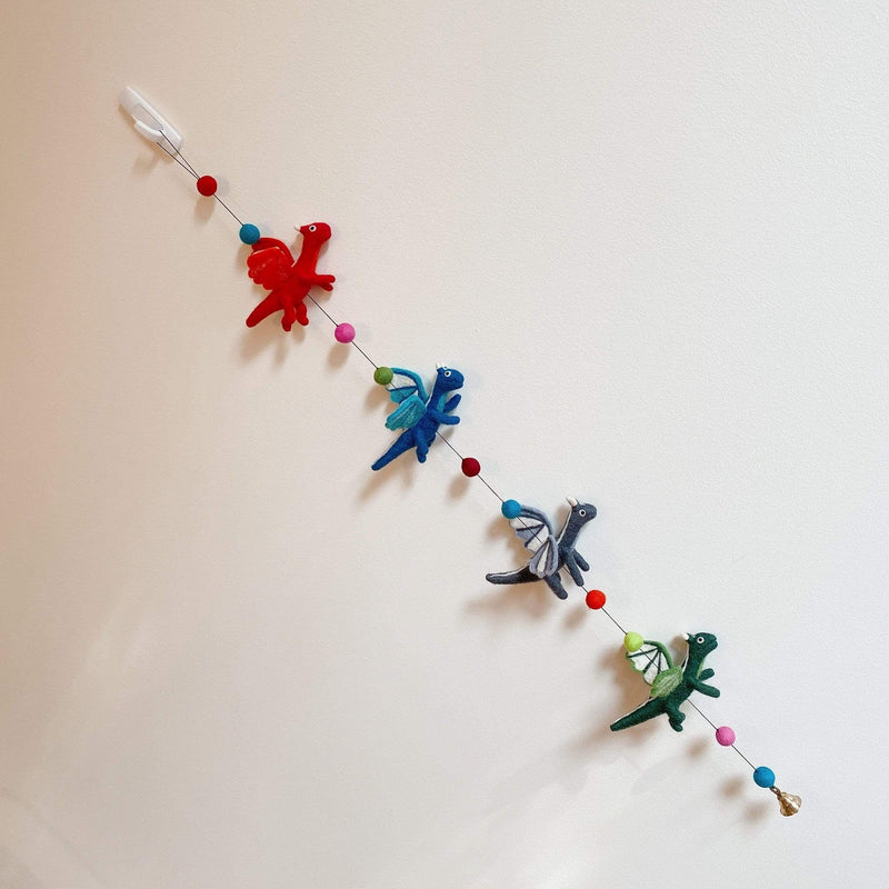 A wool felt dragon garland hangs against a light wall, featuring four 3D felted pterosaurs. These playful dragons are red, blue, gray, and green, strung along the approximately 4-foot long garland. A small copper bell dangles from one end, adding a whimsical touch.