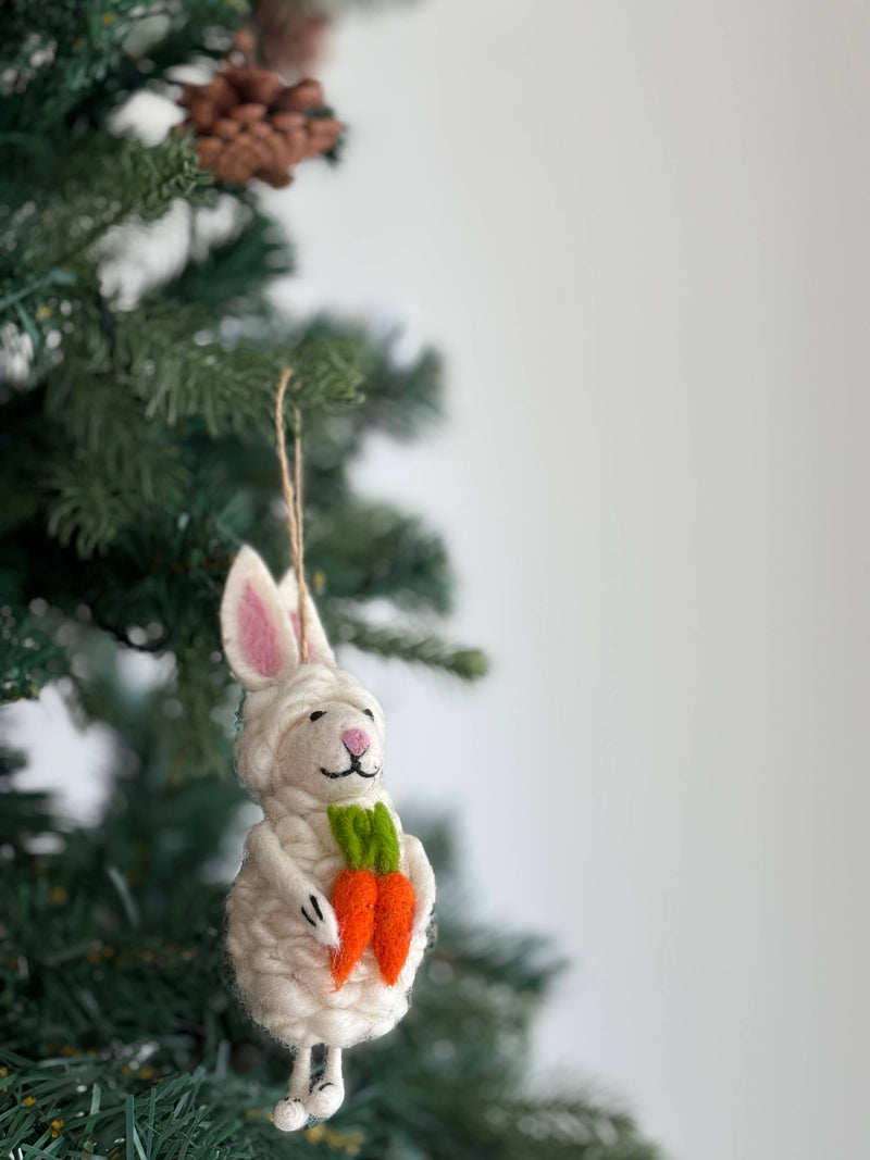 A cream-colored Felt Easter Bunny Sheep Ornament made of wool felt hangs on a Christmas tree. This unique felted ornament features a fluffy sheep with long bunny ears that have pink insides, a stitched pink nose, and a black stitched smile. The sheep ornament charmingly holds two orange felted carrots with green tops, with a jute string visible for hanging.