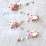 A colorful felt garland featuring four flying pig ornaments, each adorned with pink pigs, white pom-poms, and wings, hanging from a string against a neutral marble countertop background. The whimsical design exudes a playful and festive atmosphere, perfect for decorating a child's room or celebrating special occasions.