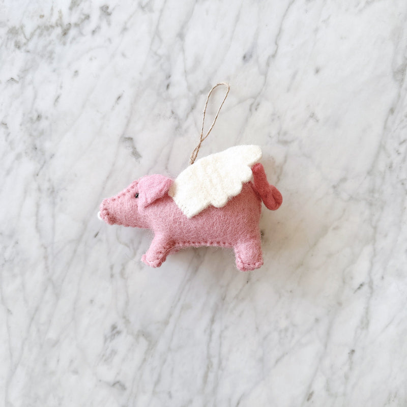 A handcrafted wool felt flying pig garland hangs against a marble backdrop, featuring multiple pink pigs with white wings attached. The neutral background provides a contrasting setting for the whimsical garland. Each pig's soft and durable wool or felt wings give the impression of soaring through the air.