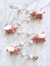 A collection of felt flying pig garlands rests on a marble countertop, each featuring a pink pig with wings attached to its back. The pigs are connected by strings, suspended in mid-air, giving the impression that they're flying. Made from soft wool felt, these whimsical garlands have a cute design and a soft texture.