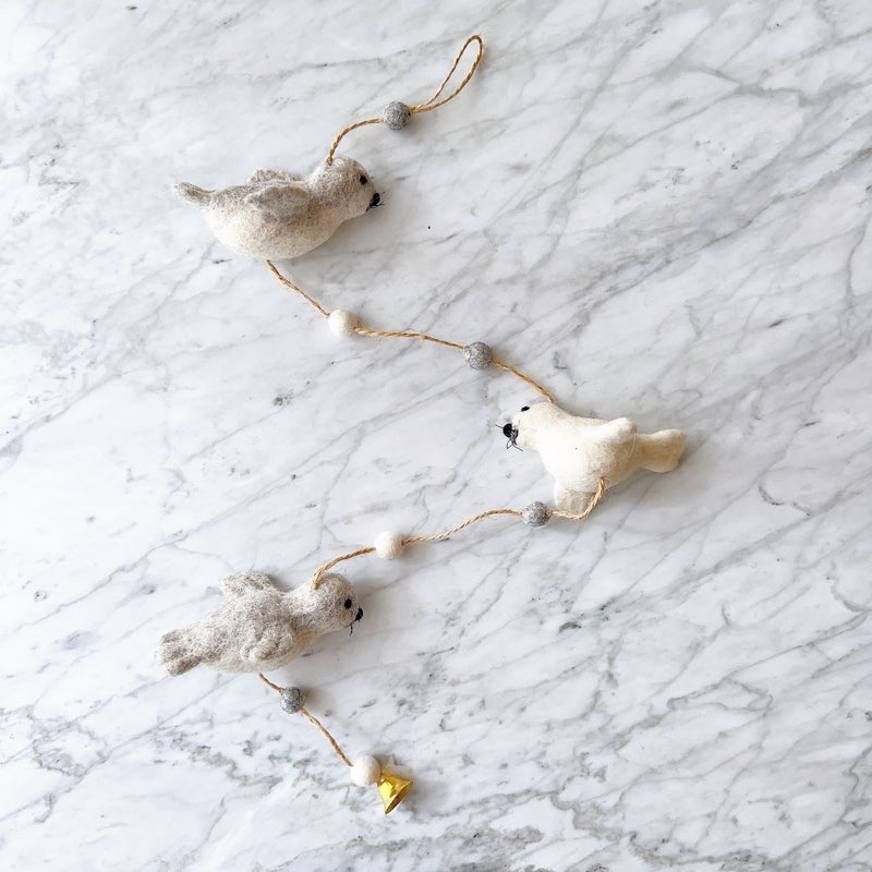 A felt seal garland is displayed on a white marble surface, exuding a calm and playful atmosphere. The garland features three soft felt seals—two light grey and one white—interspersed with alternating white and grey felt balls on a brown string. The bottom felt seal is adorned with a small copper bell.
