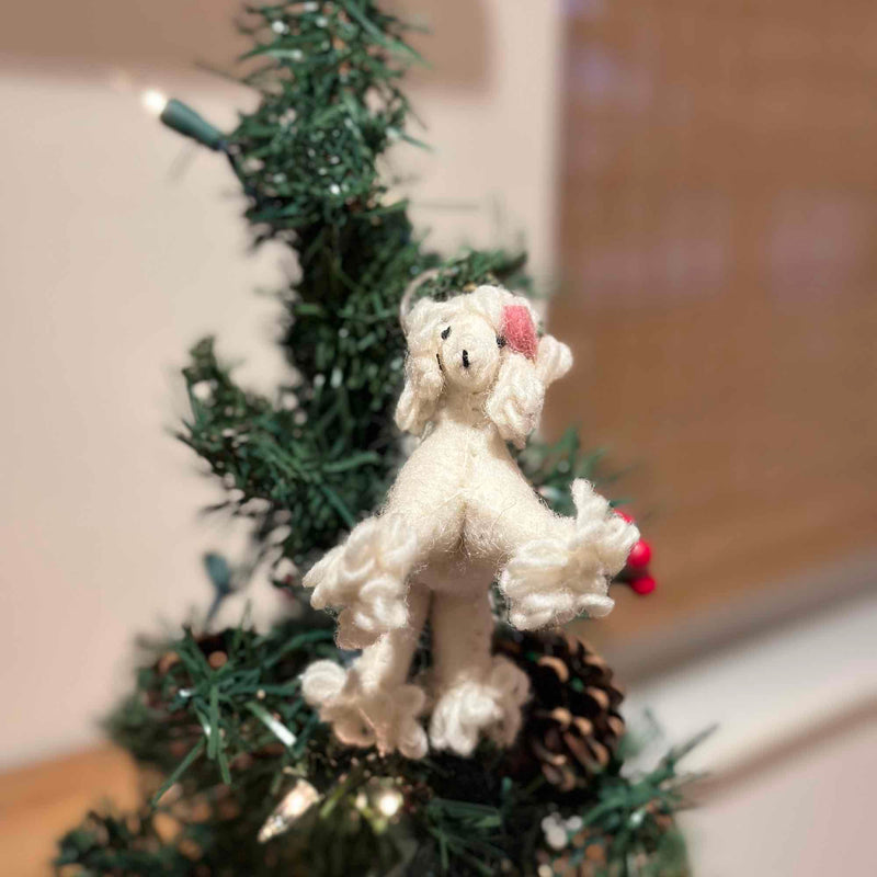 A charming, handcrafted white poodle felt keychain hangs on a festive green Christmas tree, nestled among pine needles and shiny ornaments. Crafted from wool felt, the poodle features curly white fur, small black eyes, and a delicate pink bow on its head, adding to its cozy seasonal aesthetic.