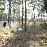 A wool felt mountain and cloud garland is strung between two trees in a sun-dappled forest. This handcrafted garland features alternating 3D felt balls, felt trees, felt clouds, and felt mountains, creating a serene, natural scene.